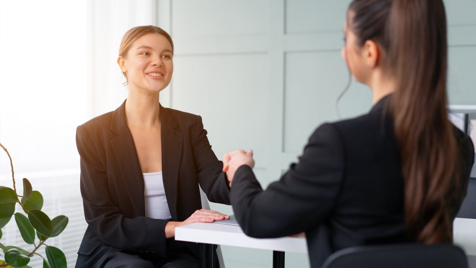 Two professional women shaking hands across a desk at a job interview